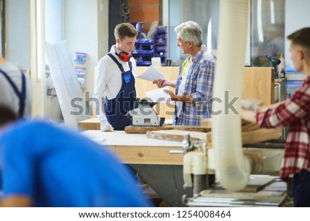 Irritated emotional senior foreman standing at workbench with polish machine and yelling at inexperienced carpenter for mistakes in sketches in factory workshop