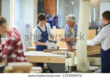Serious skilled senior carpenter with gray hair standing at desk and gesturing hand while teaching young worker to use polish machine at factory