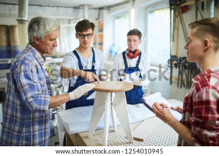 Curious carpentry students asking questions at practical class on furniture production: handsome young man in glasses pointing at stool while talking to professional carpenter