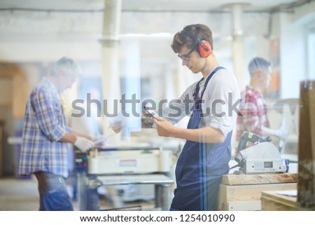 Serious busy young joiner in blue overall and ear protectors leaning on workbench with circular saw and reading operation manual in clipboard