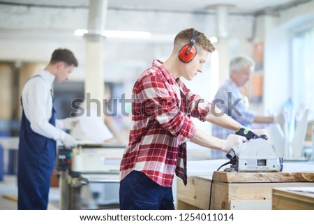 Serious concentrated young male carpenter in ear protectors standing at desk and working with circular saw while cutting wooden plank in workshop