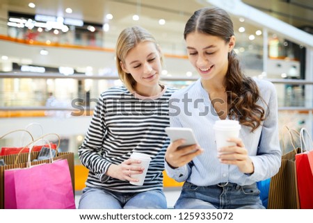 Two girls with drinks searching in online shops and comparing prices with those in offline departments