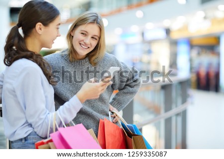 One of young shoppers looking at her friend showing curious sale notification in smartphone after shopping in the mall