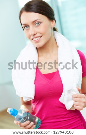 Portrait of pretty girl with towel and bottle of water looking at camera in gym