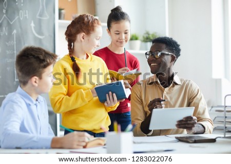 Group of pupils with books or dictionaries and pencils standing by their teacher with tablet while discussing new words