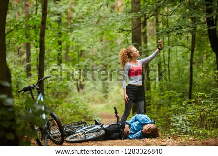 Young female cyclist with smartphone trying to catch signal in the forest while standing on path by her boyfriend with broken leg lying on the ground.