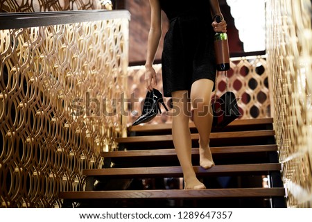 Drunk young woman in black dress holding her shoes, handbag and bottle of wine while walking downstairs inside restaurant