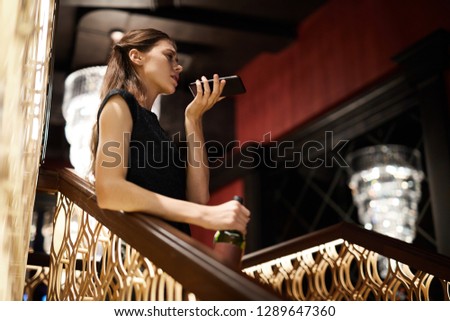 Drunk girl with bottle of wine recording voice message on smartphone while leaning against railings in restaurant