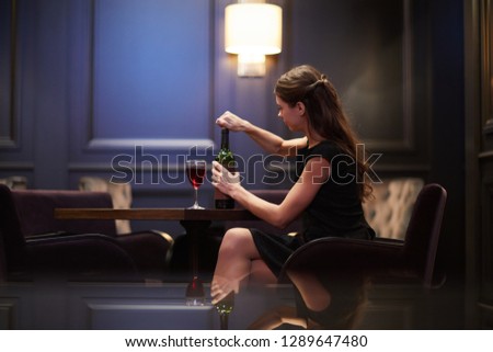Young woman opening or covering bottle of red wine while sitting by table in classy restaurant alone