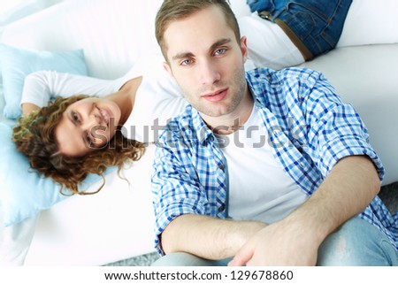 Young man sitting on the floor and looking at camera with his girlfriend lying on sofa on background