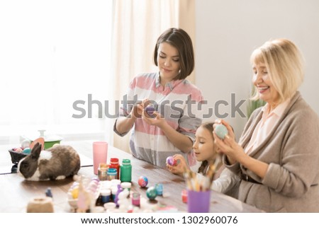 Happy women and little girl painting eggs for Easter by table with cute small rabbit near by