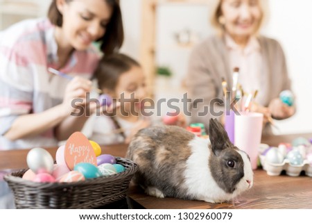 Cute fluffy rabbit sitting on wooden table by basket with Easter eggs being painted for holiday by two women and little girl