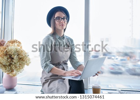 Young owner of florist studio with laptop working with online orders while sitting by window