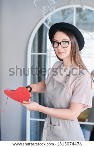 Pretty girl in apron and hat cutting red paper heart while standing in front of camera in studio