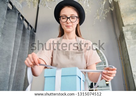 Young female tying up silk ribbon on top of blue giftbox while working in studio or shop