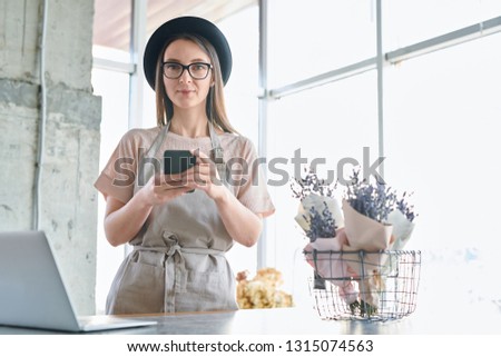 Young woman in workwear, eyeglasses and hat scrolling in smartphone while working in floral studio