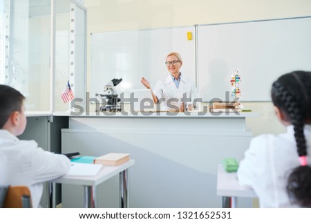 Aged blonde teacher of chemistry pointing at one of schoolkids while asking him question at lesson