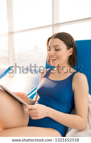 Happy young mobile female in blue swimsuit relaxing on deckchair and watching online video