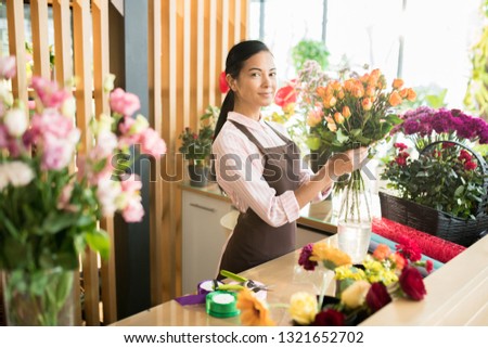 Young female florist in apron and shirt standing by table with supplies and flowers while preparing new bouquet for client
