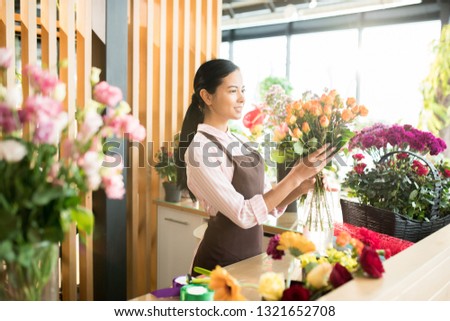 Pretty brunette girl in apron looking at big rose bouquet in her hands while making floral bouquets in florist shop