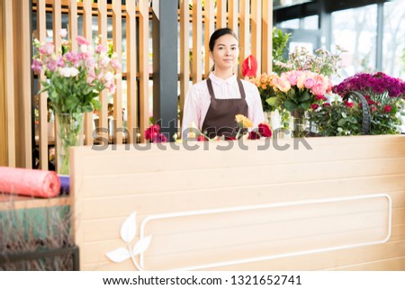 Young friendly florist shop assistant in apron standing by counter and waiting for new clients