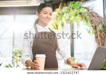 Happy young florist in apron having drink at break and looking through online data in laptop