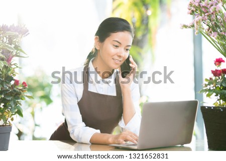 Young brunette female in apron searching for online information in front of laptop and talking to client on the phone