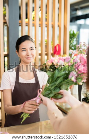 Happy young woman in apron selling fresh flowers to buyers and clients of her own floral shop
