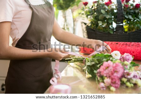 Florist in apron tying nude pink silk ribbon aroung fresh floral bouquet consisting of various flowers