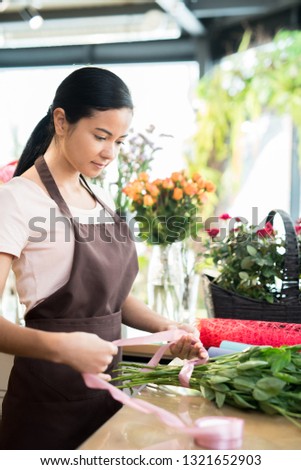 Young woman in apron tying bunch of fresh flowers with pastel pink silk ribbon while working in florist shop