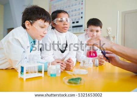 Secondary school students in whitecoats and eyeglasses looking at liquid substance in tube and listening to explanations of teacher