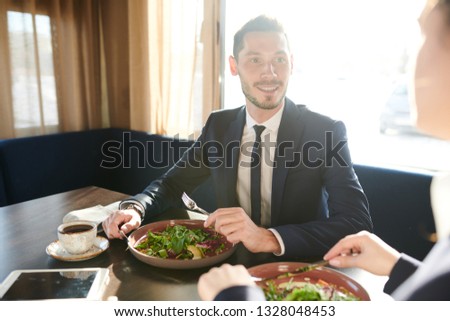 Young confident man in elegant suit talking at colleague while having fresh salad for lunch in cafe or restaurant