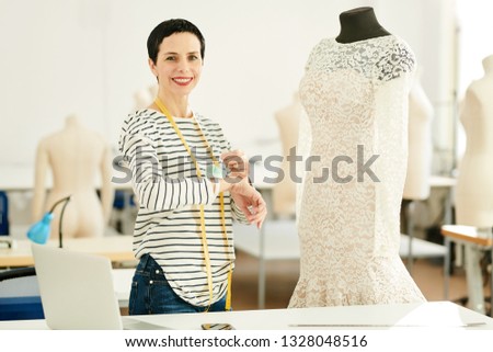 Happy brunette professional tailor and fashion designer standing in her studio or workshop by dummy