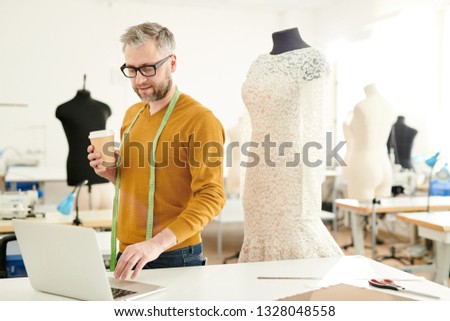 Mature professional fashion designer with measuring tape and glass of drink standing by desk in front of laptop in studio