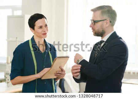 Happy female tailor having conversation with one of clients and specifying details in notepad