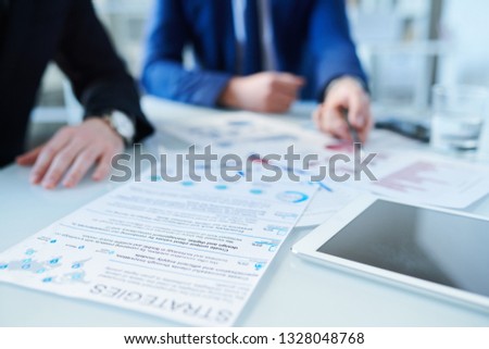 Paper with list of business strategies, other documents and touchpad on desk with two working businessmen on background