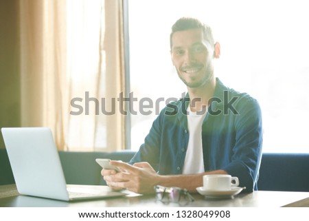 Young cheerful businessman with smartphone sitting by table in front of laptop and texting at work