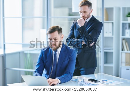 Pensive businessman in suit standing behind colleague sitting by desk in front of laptop and both looking at display