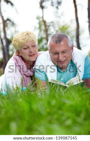 Happy mature couple lying on grass and reading a book in the countryside