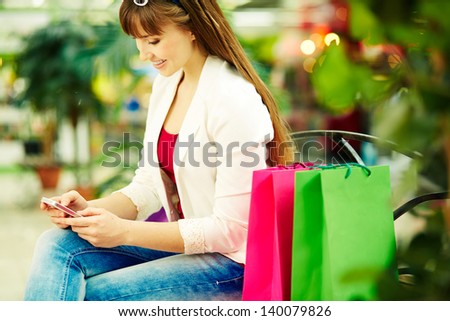 Pretty lady with colorful shopping bags sitting in trade center and writing sms