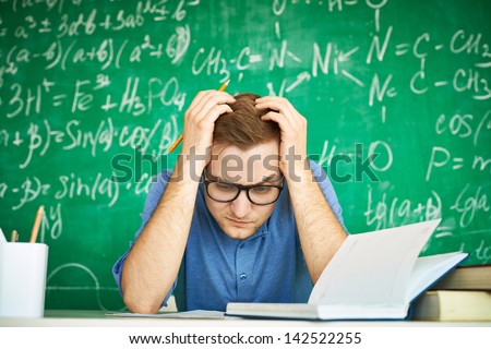 Portrait of teenage student with his head in hands carrying out graduation test on background of chalkboard