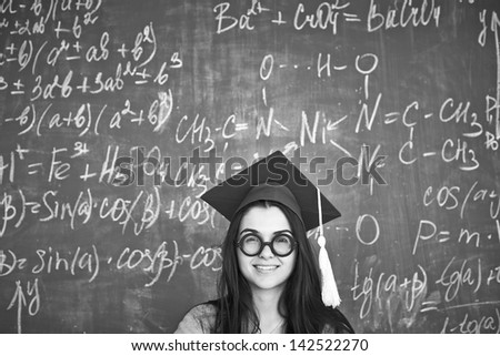 Black-and-white image of student in graduation hat and eyeglasses looking at camera on background of chalkboard