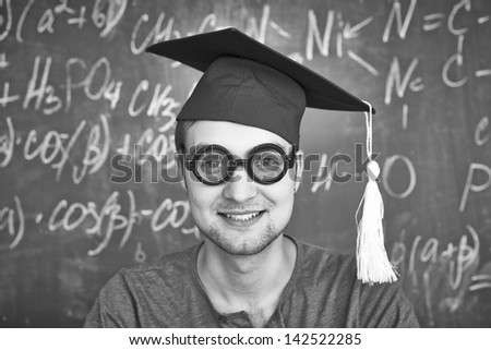 Black-and-white image of handsome student in graduation hat and eyeglasses looking at camera on background of chalkboard