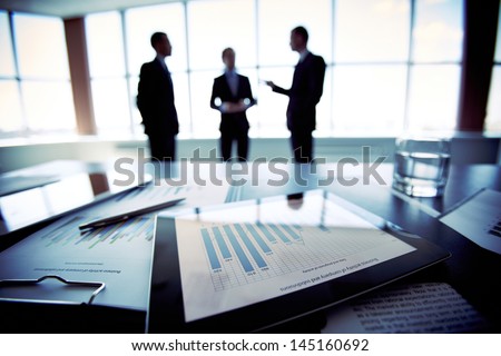 Close-Shot Of A Tablet Computer Displaying Financial Data, Three Businessmen Standing In The Background