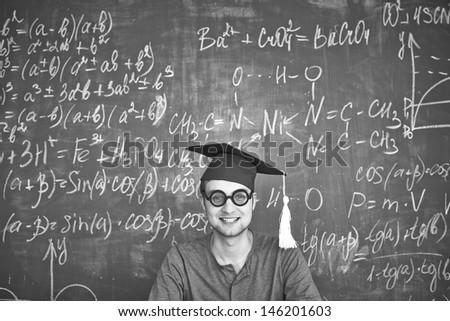 Black-and-white image of student in graduation hat and eyeglasses looking at camera on background of chalkboard