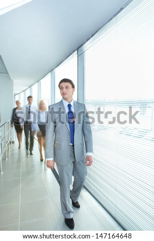 Portrait of confident businessman walking along corridor with team of partners on background