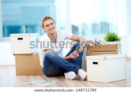 A young guy sitting on the floor of new house surrounded with boxes and choosing color from palette