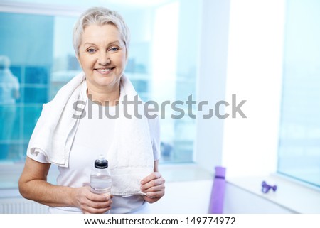 Portrait of pretty senior woman with towel and bottle of water in gym