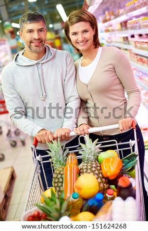 Image of happy couple with cart full of products looking at camera in supermarket