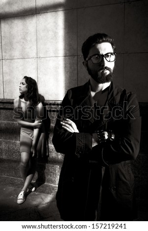 Black-and-white image of calm man with crossed arms on background of young female standing by wall